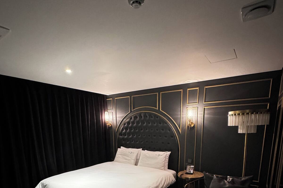 A modern bedroom with a large bed, a round side table, a small round ottoman, and a wall-mounted lamp.