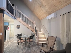 Un escalier mène à une chambre à coucher située en mezzanine, avec un plafond en bois. On aperçoit également un coin repas avec une table et des chaises, ainsi qu’une cuisine à l’arrière-plan.