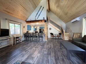 Un vaste espace de vie doté d’un haut plafond et de poutres apparentes, une cuisine avec des meubles bleus, un coin repas avec des tabourets de bar et un escalier menant à un niveau supérieur.