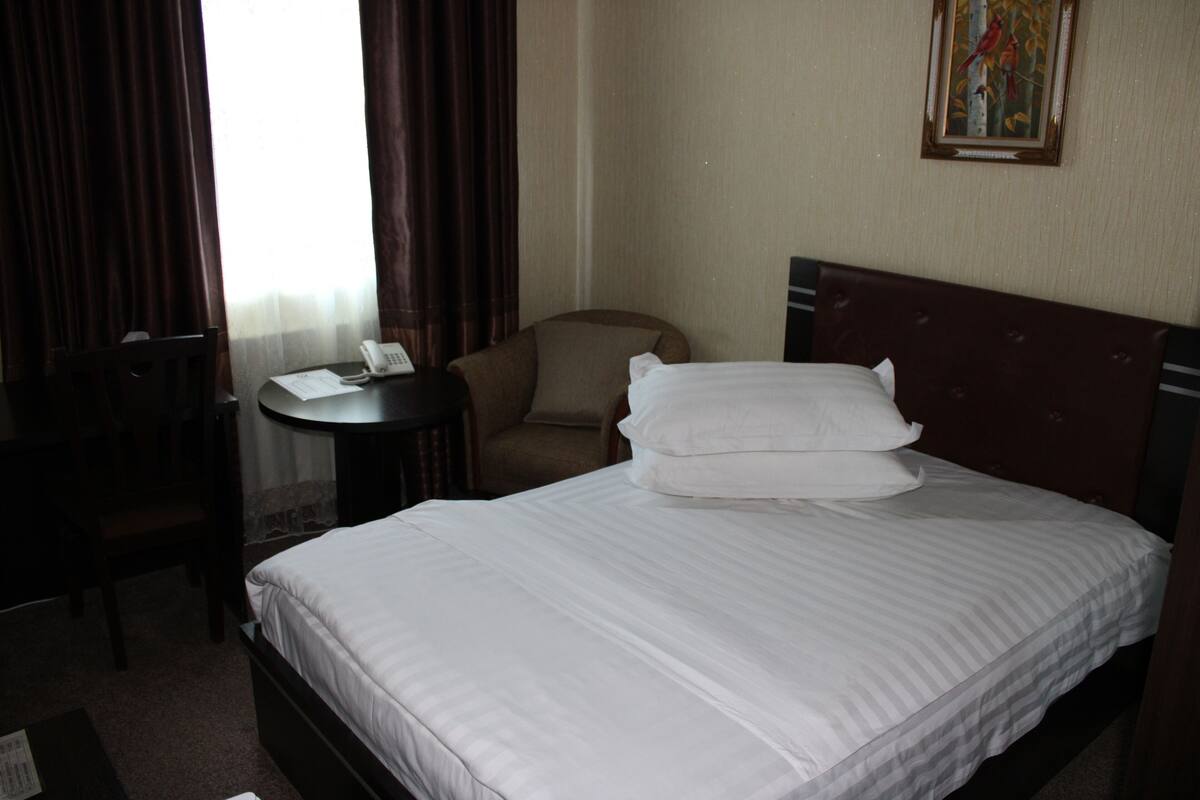 A hotel room with a bed, a chair, a small table with a phone, and a painting on the wall.