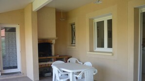 Eine überdachte Terrasse mit einem Backstein-Grill, weißen Plastikstühlen und einem Fliesenboden.