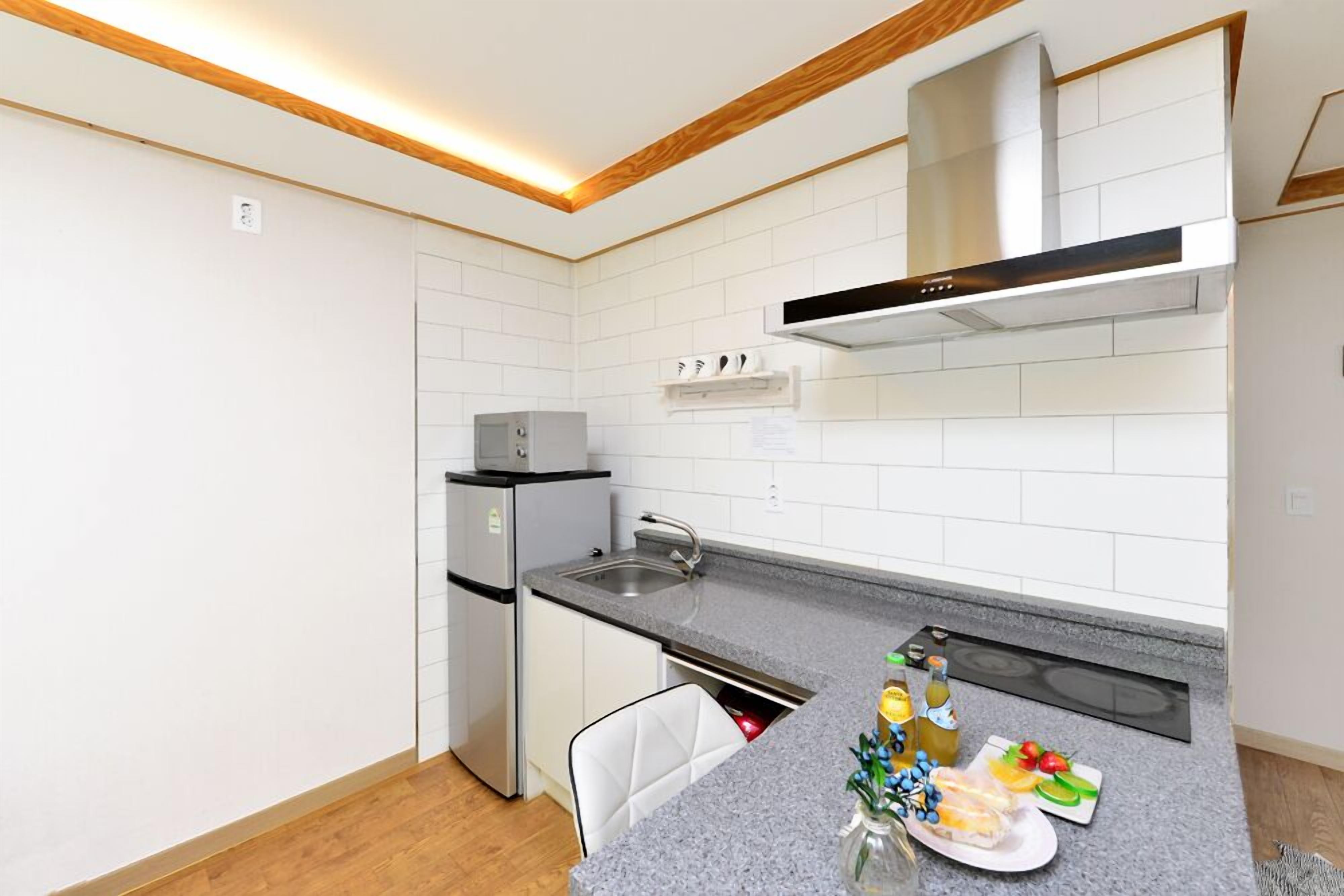 A compact kitchen with white tiles, a stainless steel range hood, a built-in oven, and a small refrigerator.