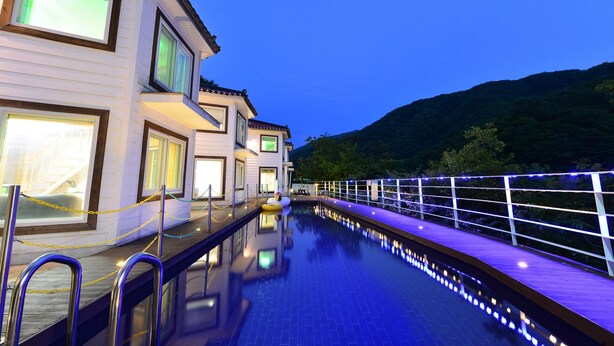 A swimming pool with a wooden deck, a white building with multiple windows, and a metal railing. - Camel Pension (Gapyeong)