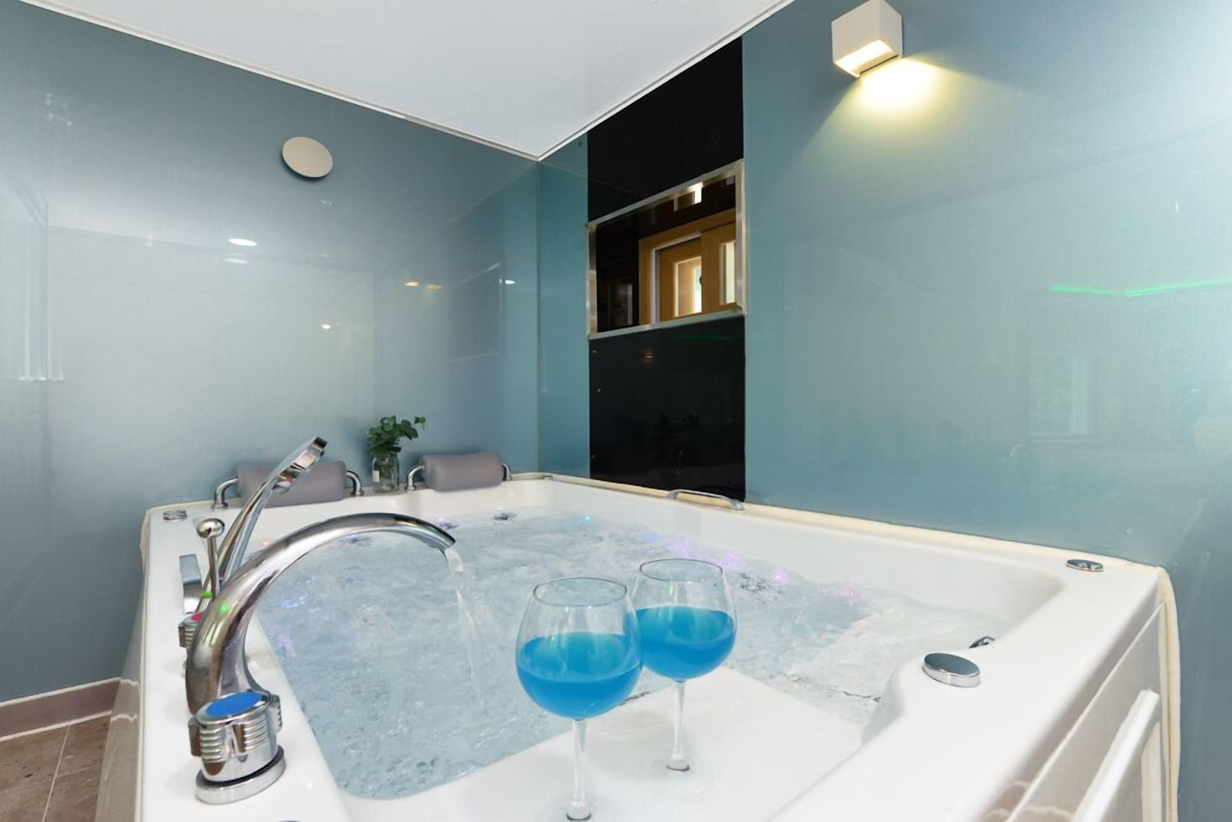 A modern bathroom with a large bathtub, two glasses of blue liquid, and a visible shower area.