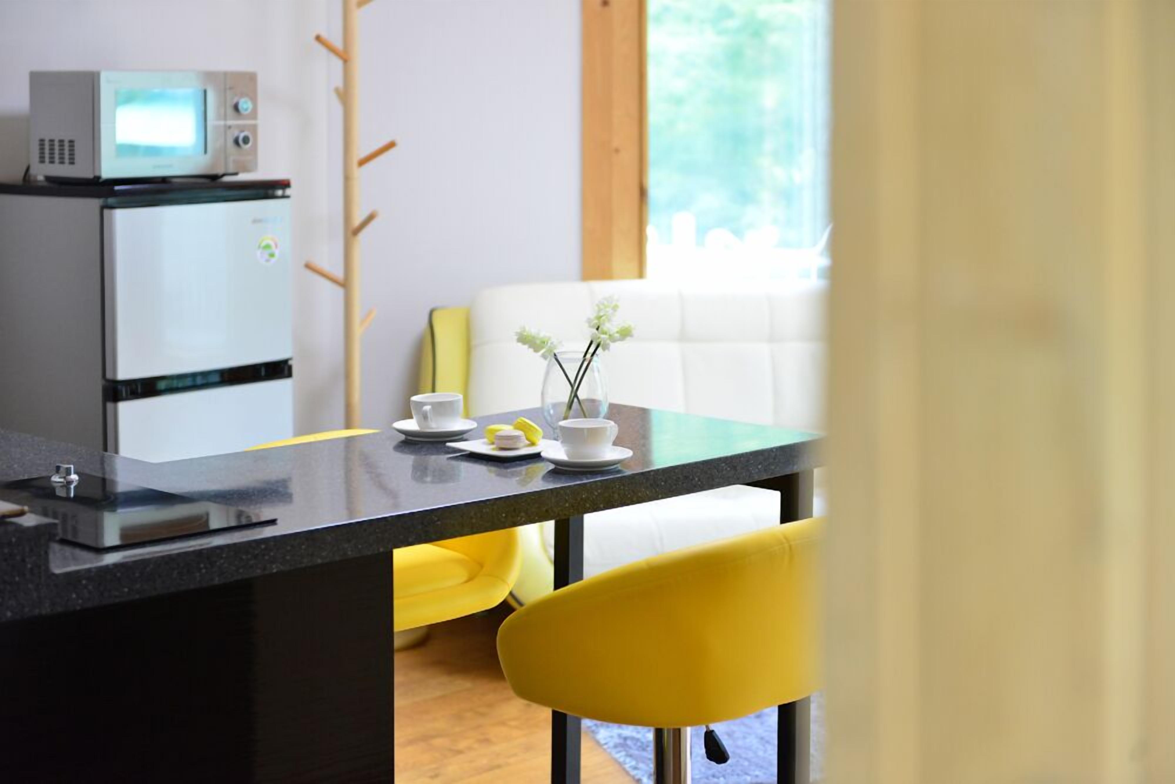 Una cocina moderna con microondas y refrigerador, una mesa de comedor con una silla amarilla y un sofá blanco al fondo.