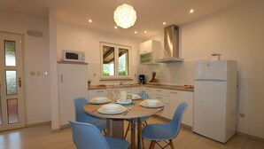 A modern kitchen with a dining table set for four, blue chairs, a white refrigerator, and a window with a view of greenery.