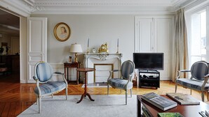 A living room with two upholstered chairs, a small round table, a fireplace with a mantel, a television, and a bookshelf. - Suburban Peace in Neuilly-Sur-Seine, by Veeve (Paris)