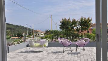 A terrace with two chairs and a planter with flowers, overlooking a hilly landscape and buildings.