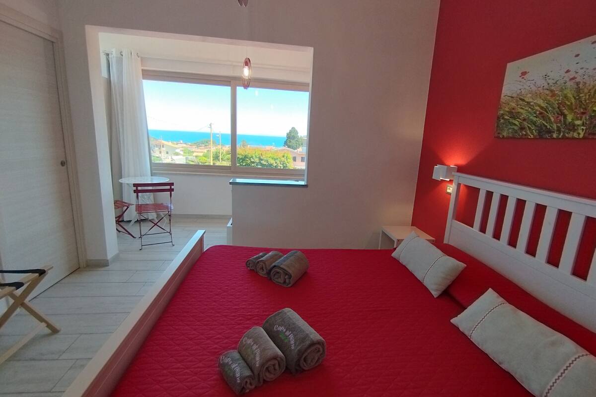 Une chambre à coucher avec une couvre-lit rouge, une tête de lit blanche et une vue sur la mer par une grande fenêtre.
