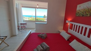 Quarto com colcha vermelha, cabeceira branca e vista para o mar por uma grande janela.