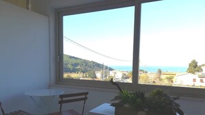 Quarto com uma janela grande que oferece vista para uma vila costeira e colinas. Há uma mesa redonda pequena e uma cadeira dobrável.