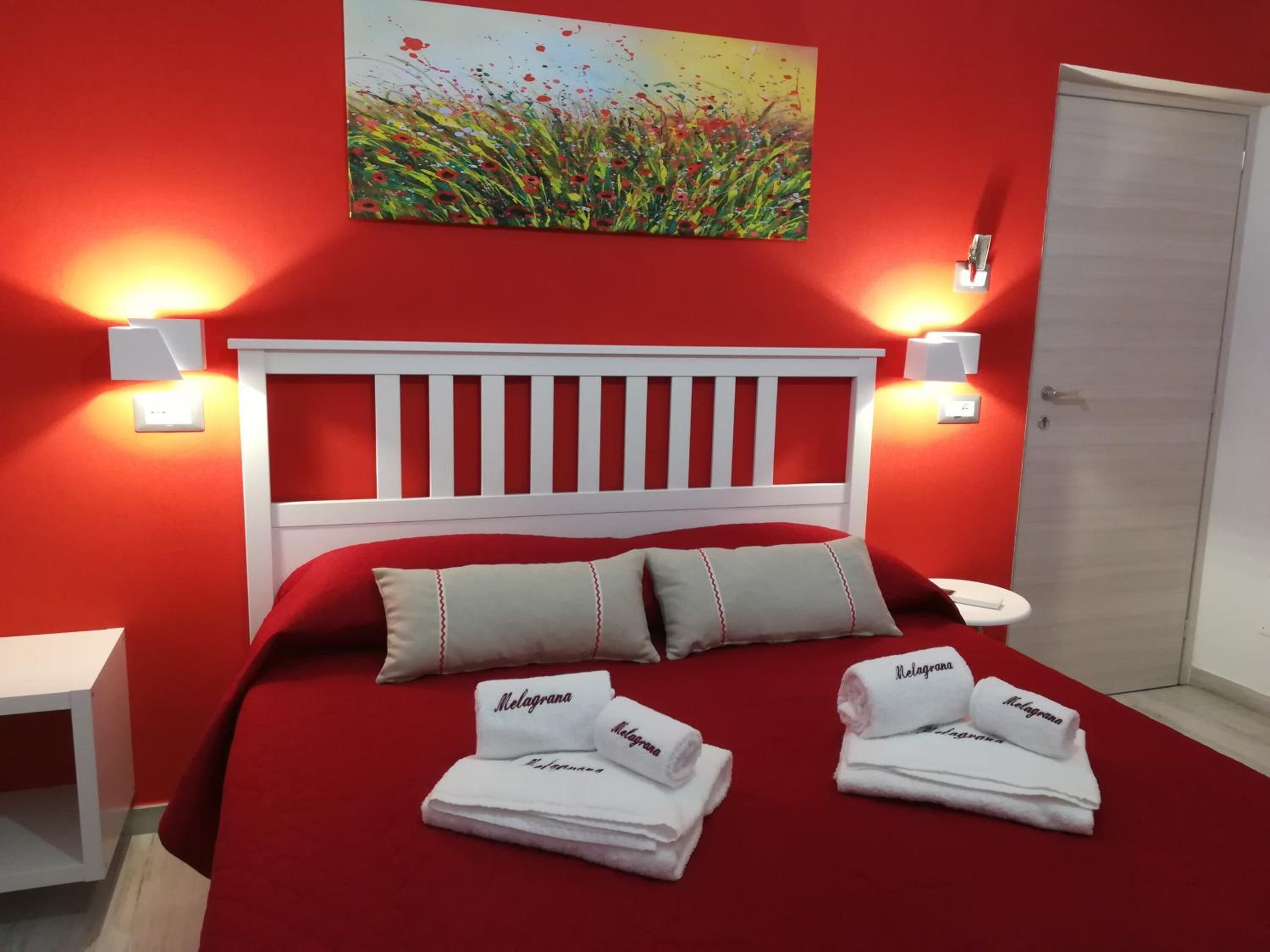 Hotelkamer met een rode muur, een wit hoofdbord en twee witte nachtkastjes. Daarboven hangt een schilderij en er liggen handdoeken met de tekst 'Melagrana' op.
