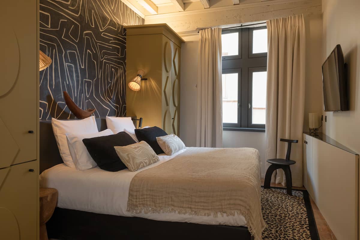A bedroom with a bed, a nightstand, a lamp, a window with curtains, and a TV mounted on the wall.