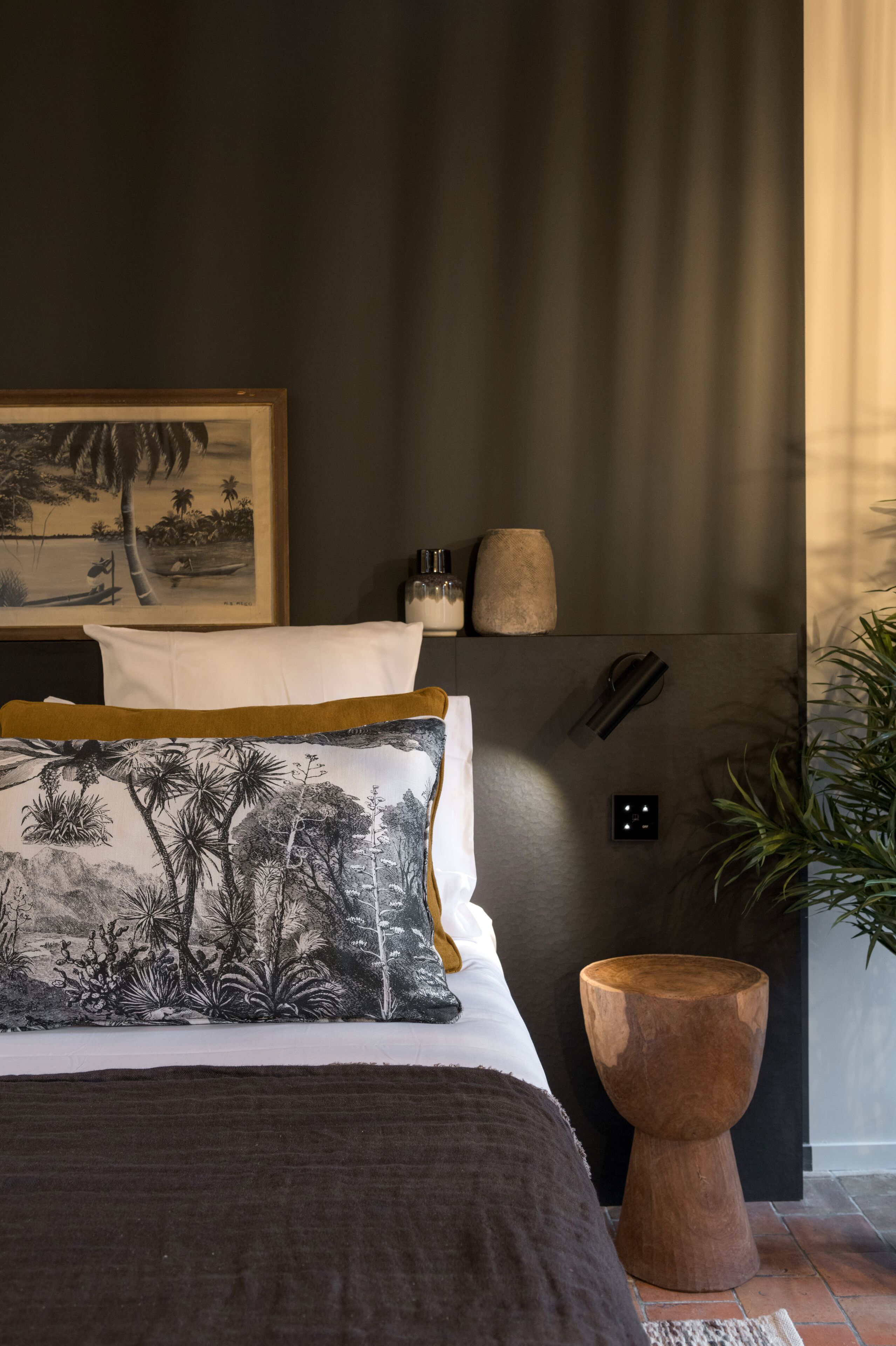 Un dormitorio con cama, almohadas, una mesita de noche, una lámpara de pared, un cuadro enmarcado y una planta en maceta.