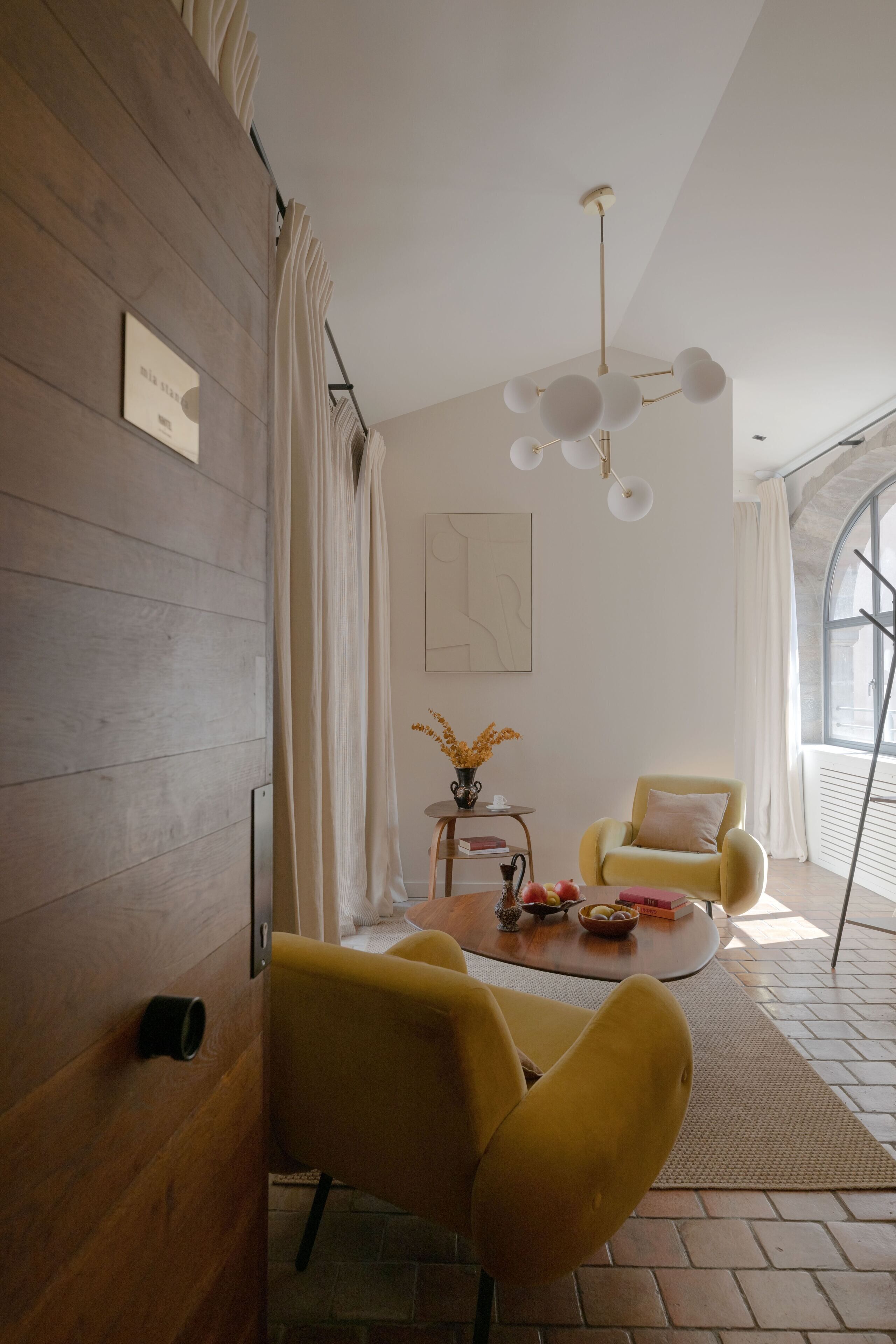 Ein modernes Zimmer mit Holzwandverkleidung, einem gelben Sessel, einem runden Couchtisch mit Dekoration, einer weißen Deckenleuchte und einem großen Fenster mit Vorhängen.