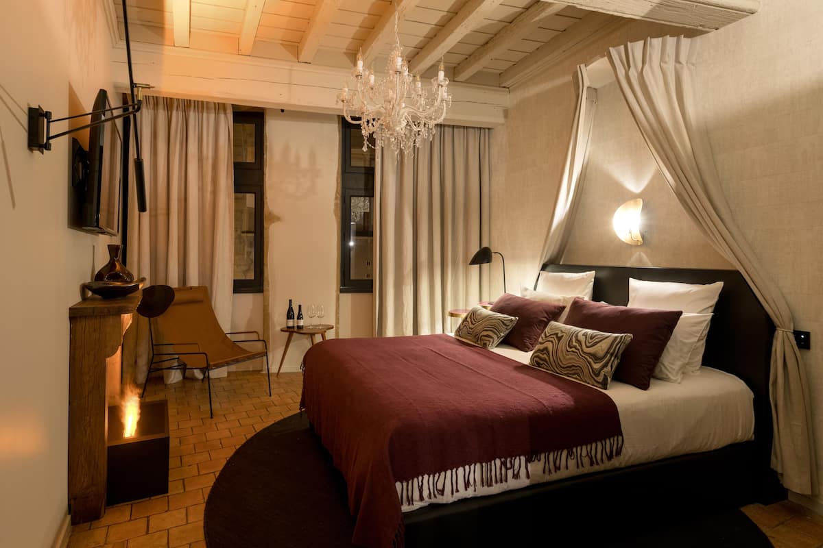 A bedroom with a large bed, a chandelier, a desk with a chair, and a view of the outside through a window.