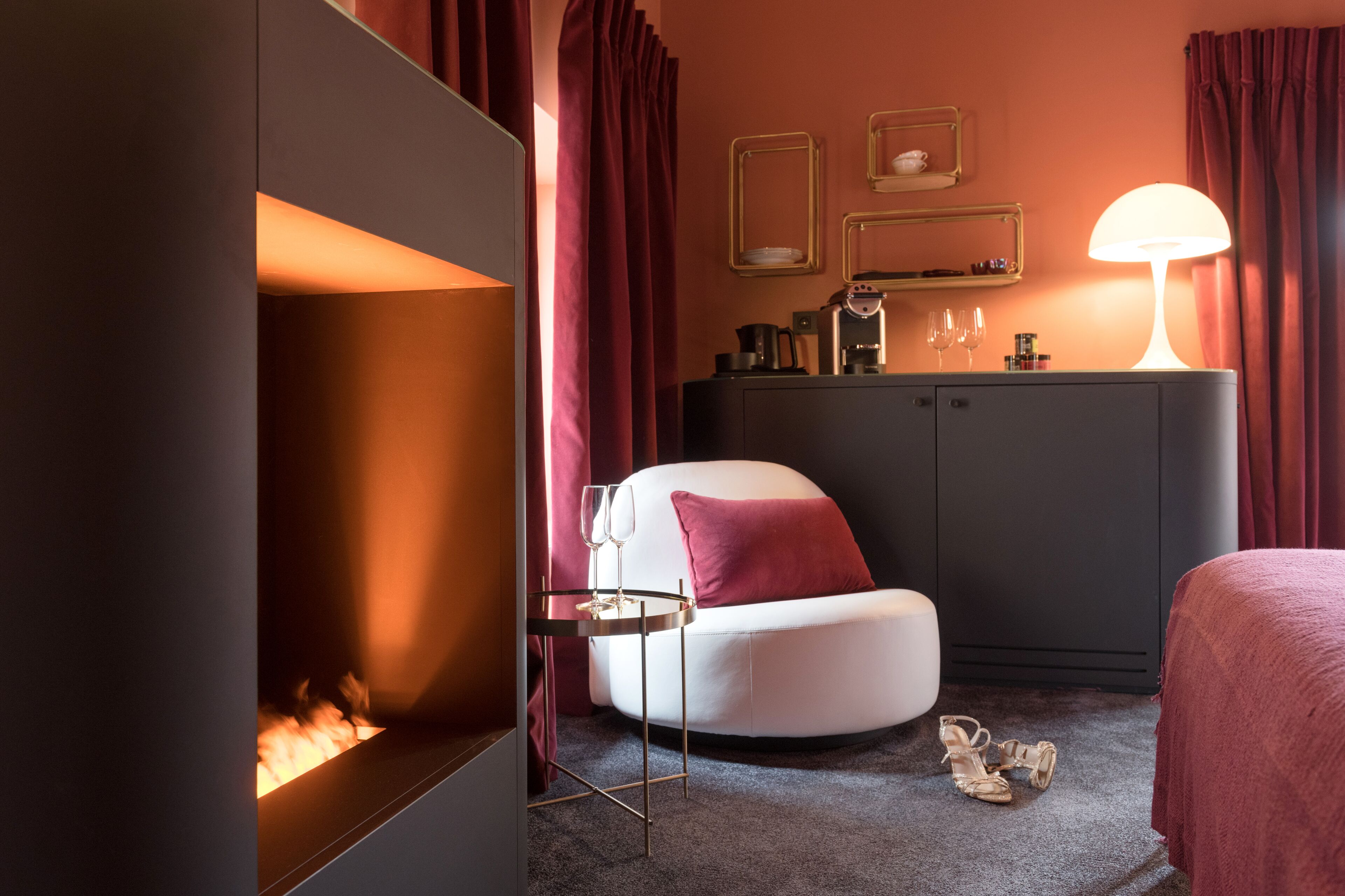 Ett modernt hotellrum med en öppen spis, en vit fåtölj, en säng med ett rött överkast, en minibar och en lampa.