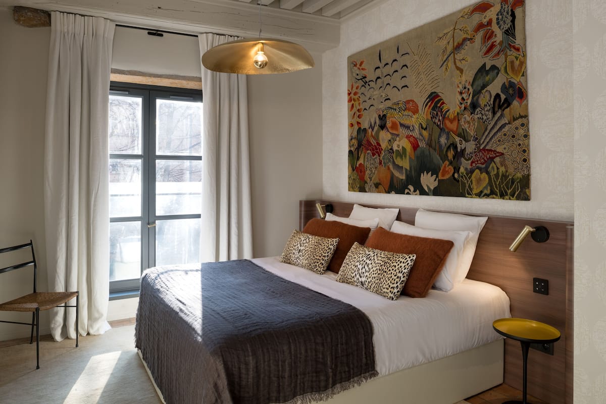 A bedroom with a large bed, a window with curtains, a painting on the wall, and a small table with a lamp.