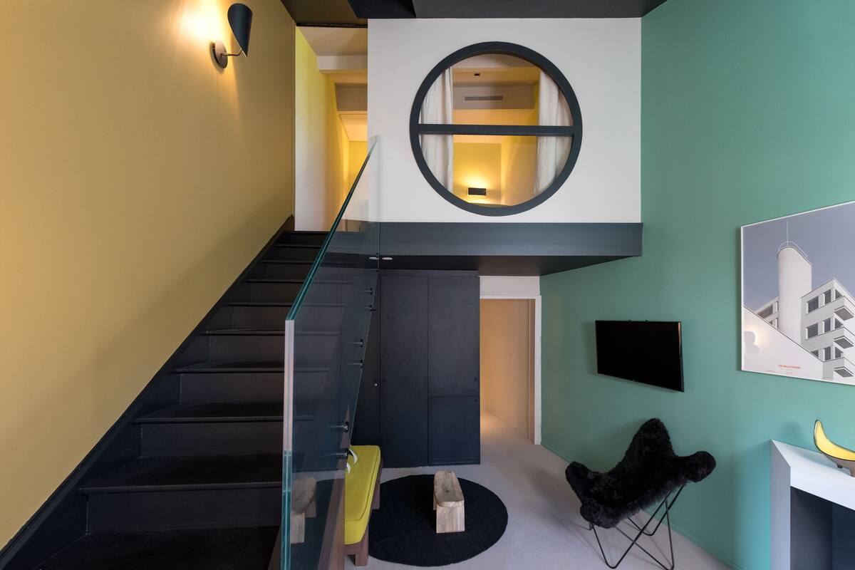 A modern interior with a staircase, a round window, a flat-screen TV, and a minimalist chair.