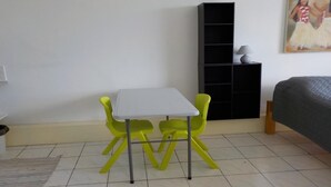 Une petite pièce avec une table blanche et deux chaises jaunes, une bibliothèque noire et un lit gris.