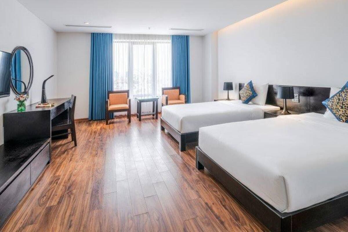 A hotel room with two beds, a desk, a chair, a TV, and a window with blue curtains.