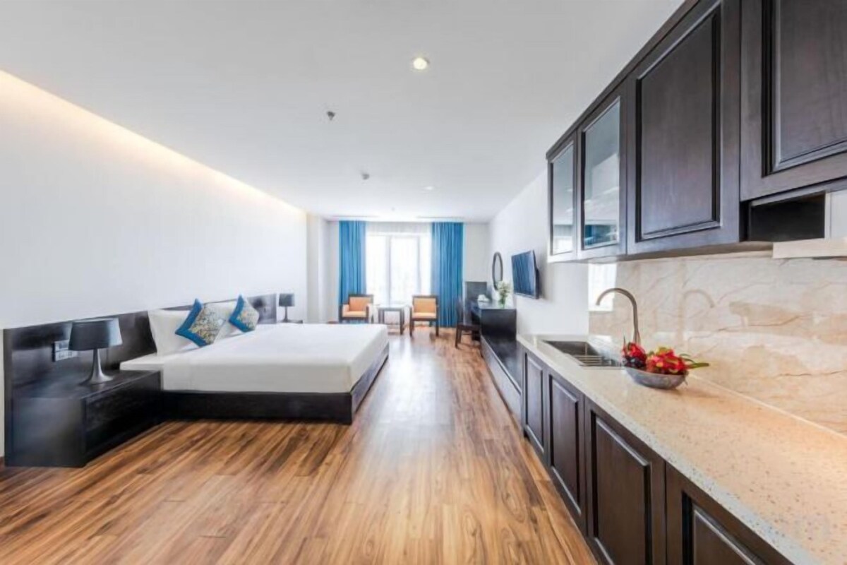 A modern hotel room with a large bed, a sitting area, a kitchen with a sink, and a view of the outdoors.