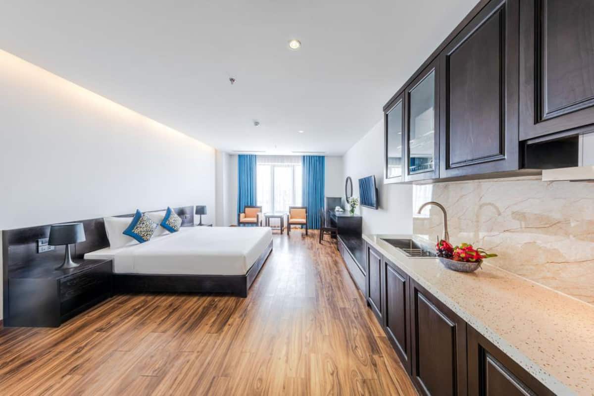 A modern hotel room with a large bed, a desk, a chair, a small table, a sink, and a mirror.