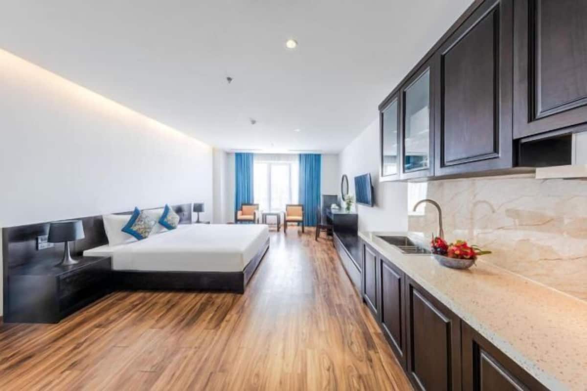 A modern hotel room with a wooden floor, a bed with a black headboard, a small desk, a chair, a TV, and a kitchen area with a sink and countertop.