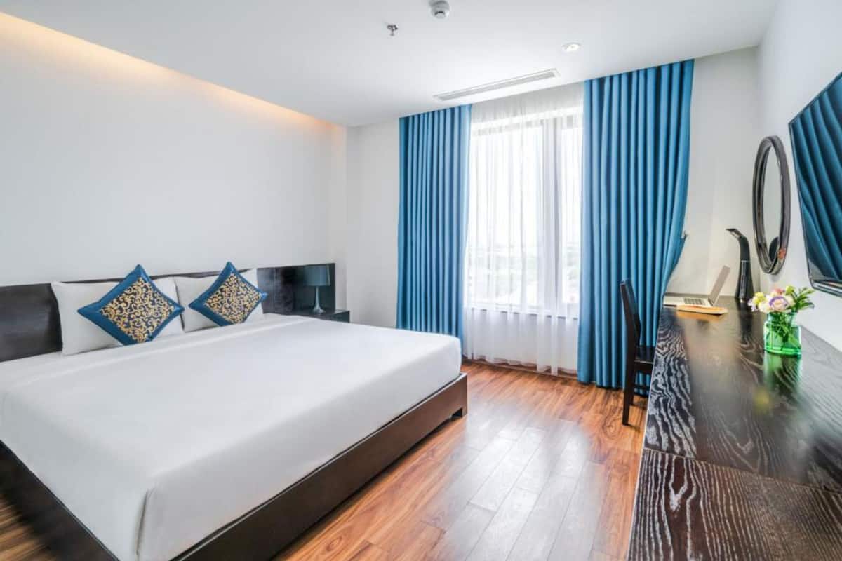 A hotel room with a large bed, a desk with a chair, a mirror, and a window with blue curtains.