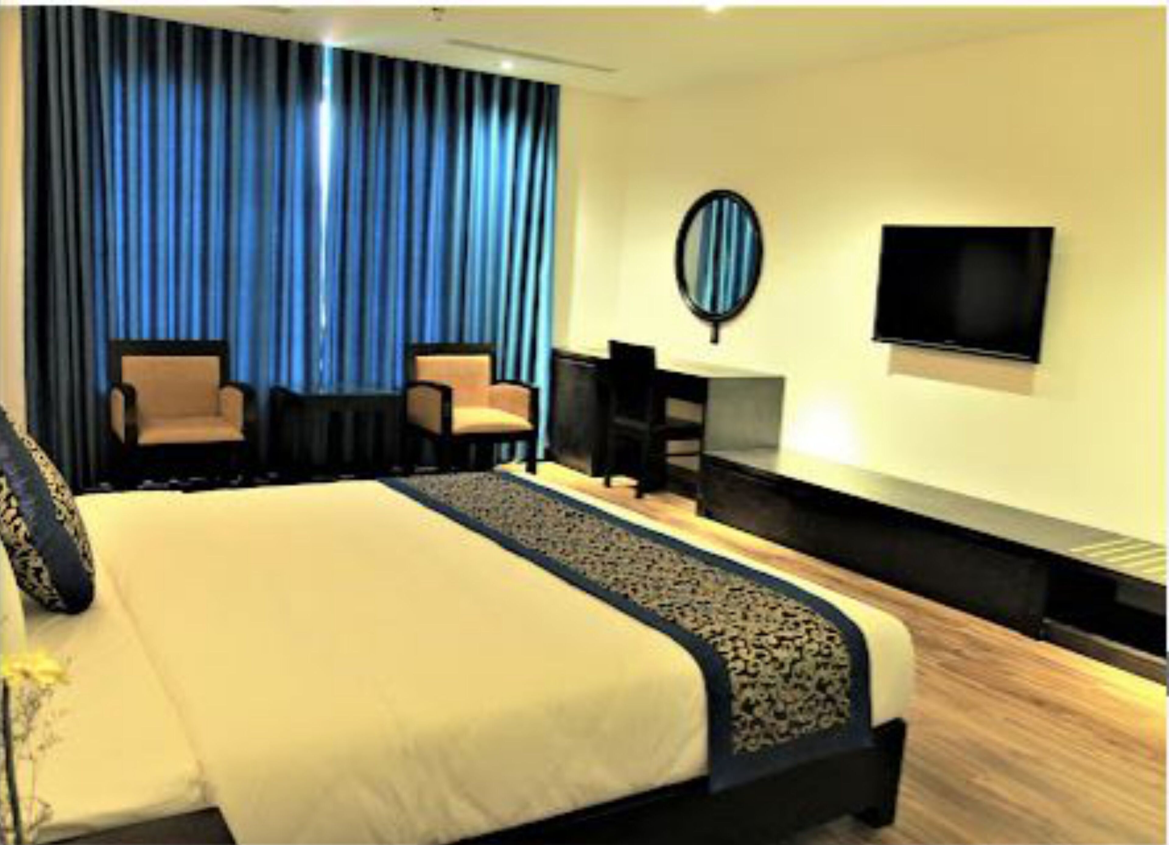 A hotel room with a large bed, two armchairs, a small table, a TV, and a window with blue curtains.