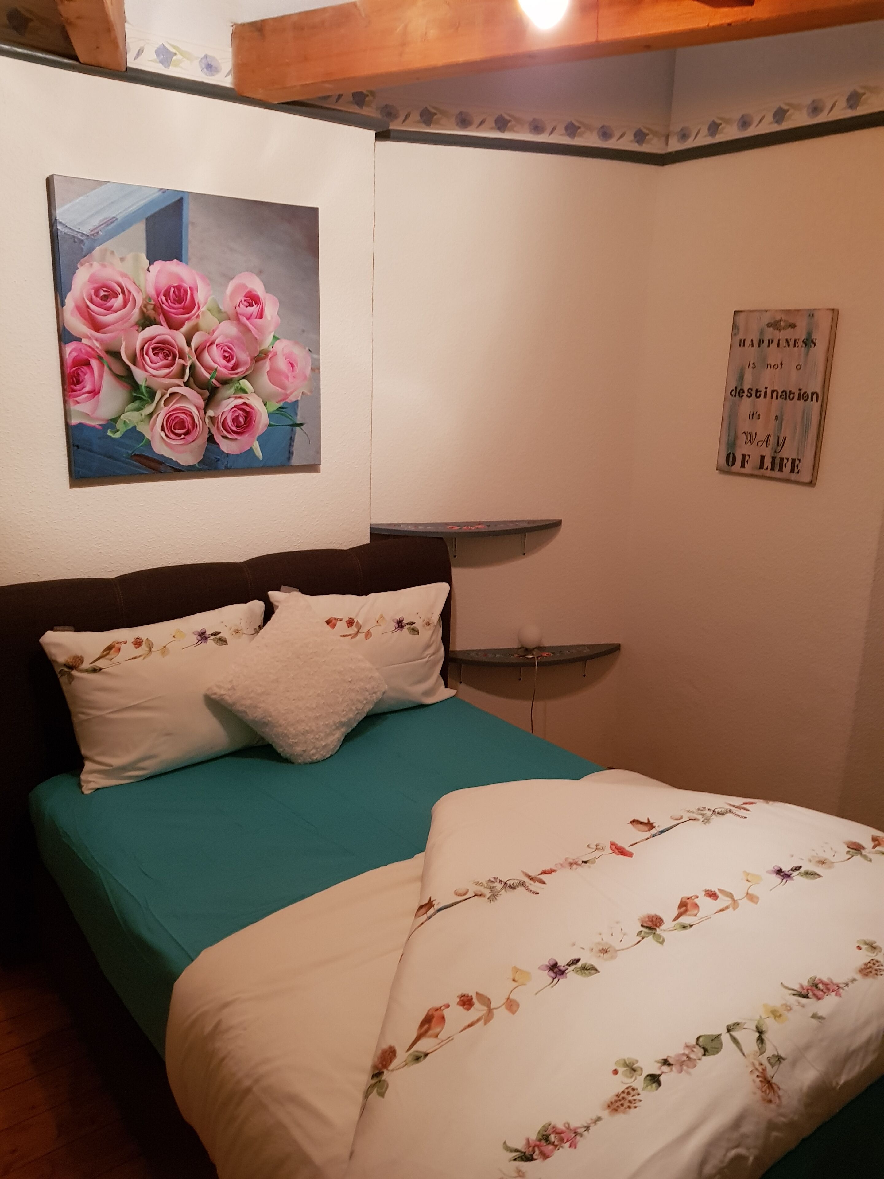 A bedroom with a bed, pillows, a wall-mounted picture of pink roses, and a motivational quote.