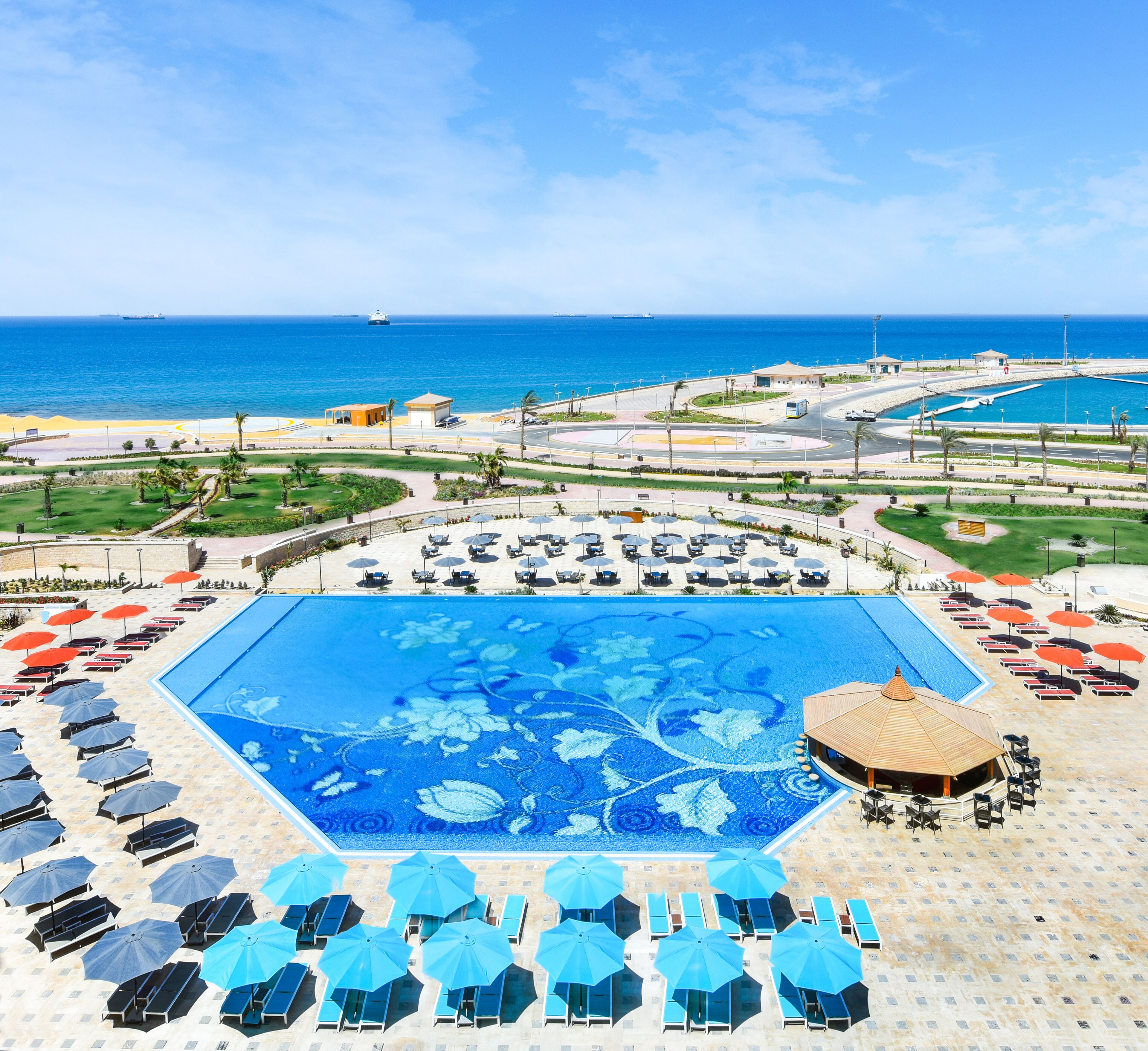 A large pool with a floral design, surrounded by lounge chairs and umbrellas, with a view of the ocean and ships in the distance.