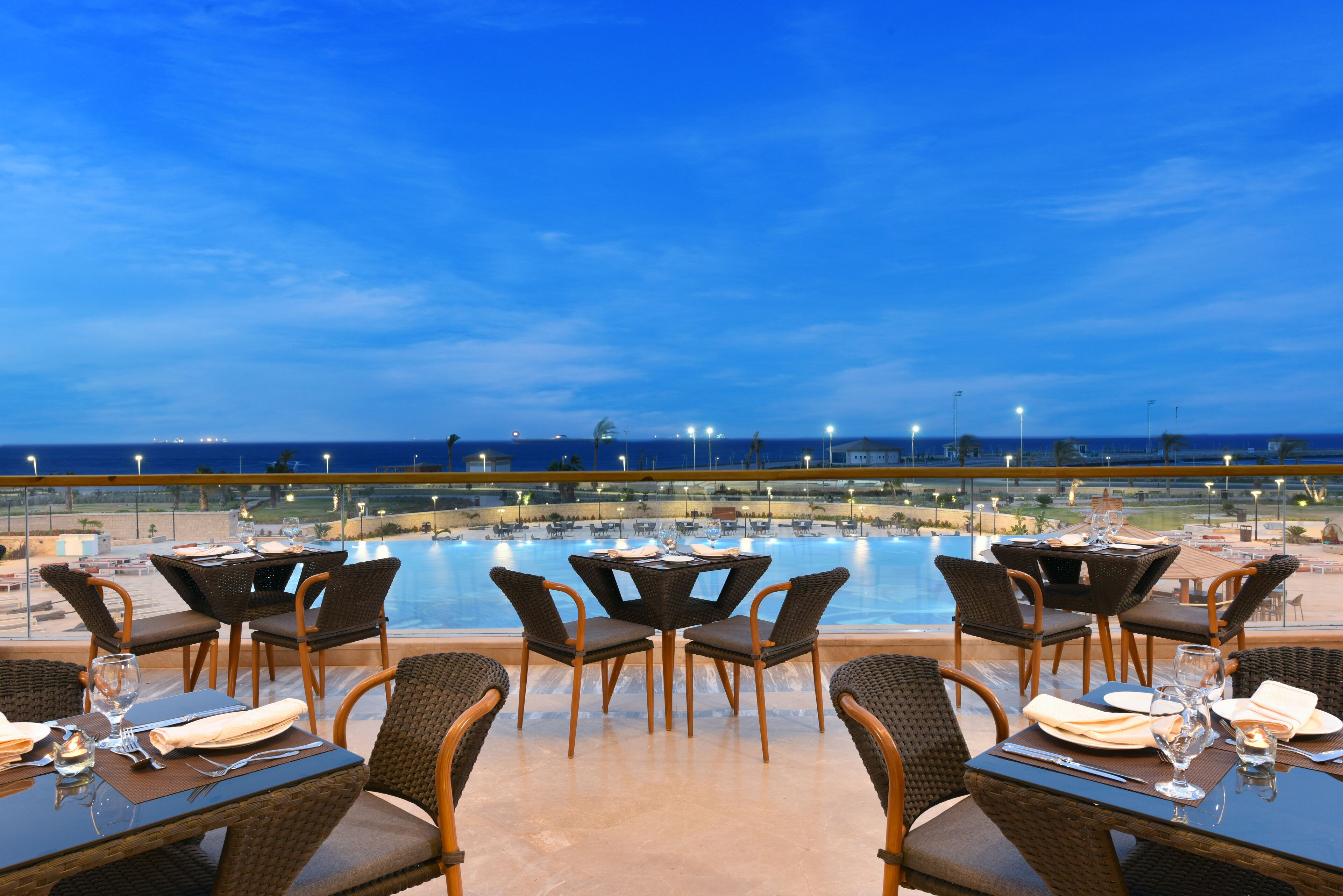 A dining area with tables and chairs overlooking a pool and the ocean at dusk.