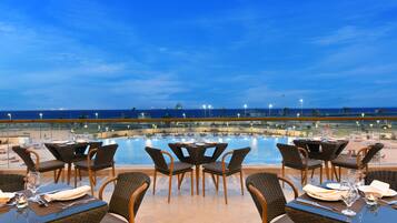 A dining area with tables and chairs overlooking a pool and the ocean at dusk.