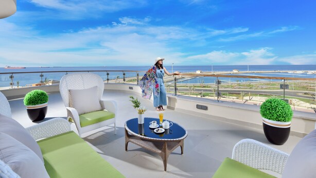 Een balkon met rieten meubels, een tafel met een drankje en uitzicht op zee en schepen.