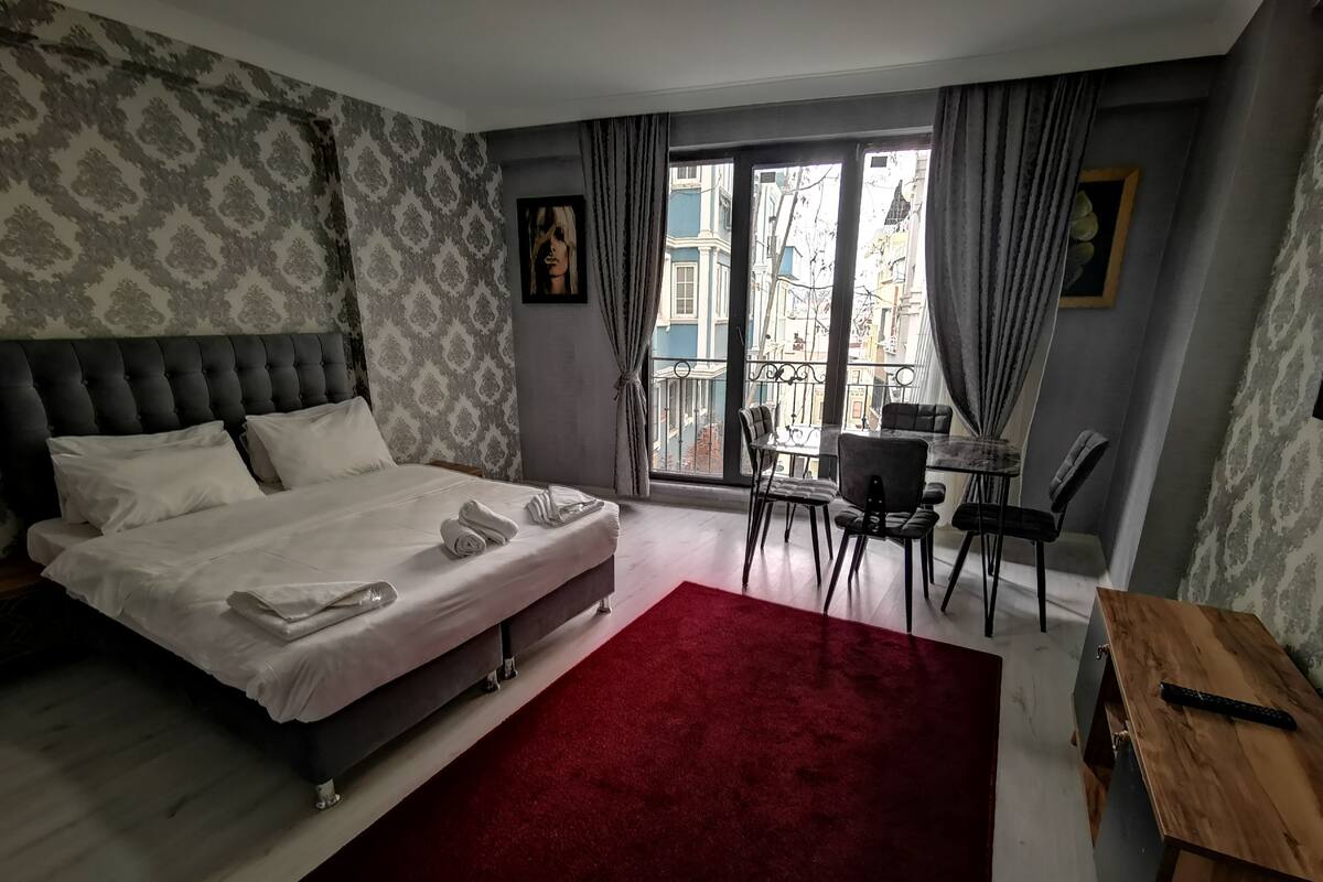 A hotel room with a large bed, a red carpet, a wooden desk, and a view of buildings through the window.