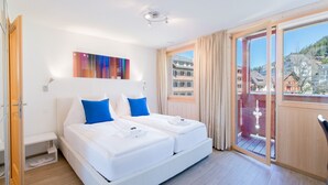 A double bed with white bedding and blue pillows, a colorful abstract painting on the wall, and a balcony with a view of buildings. - Chalet Belmont - perfect location, recently new (Wengen)
