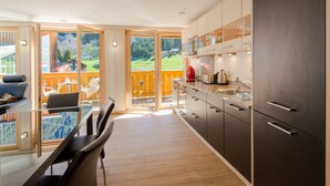 A modern kitchen with a large island, sliding glass doors to a balcony, and a view of a green landscape. - Chalet Belmont - perfect location, recently new (Wengen)
