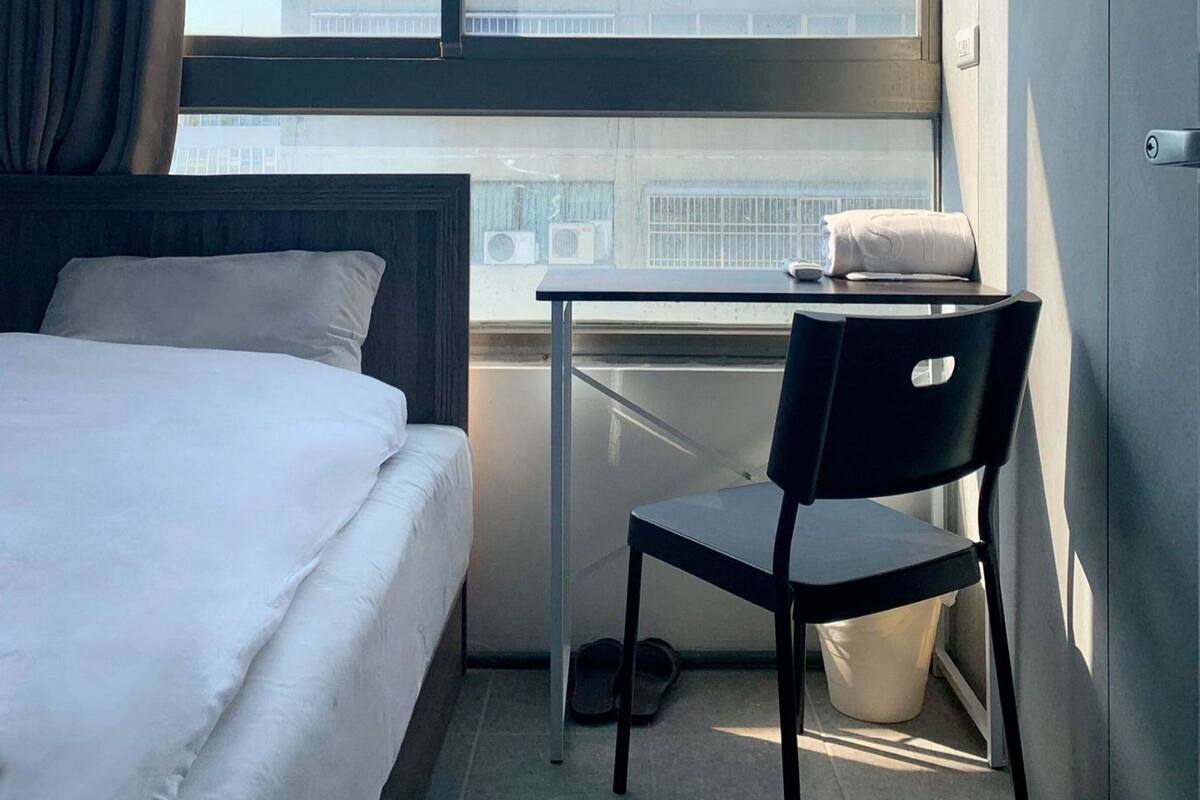 A hotel room with a bed, a desk, a chair, and a large window offering a view of buildings.