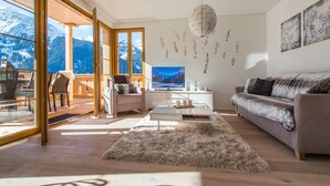 Una sala de estar moderna con sofá, mesa de centro y televisor. Hay un balcón con vistas a las montañas.