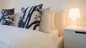 Una cama bien hecha con ropa blanca y tres almohadones decorativos con un diseño de árboles. Hay una mesita de noche con una lámpara.