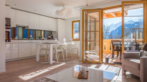 Una cocina moderna con armarios blancos, un comedor con sillas blancas y un amplio ventanal corredero que da a un balcón con vistas a la montaña.