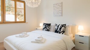 Una cama bien hecha con sábanas y toallas blancas, una ventana con vistas a árboles, y una obra de arte colgada en la pared con aves.