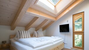 Un dormitorio con techo inclinado, una cama con ropa blanca, un televisor montado en la pared y una ventana con vistas a la vegetación.