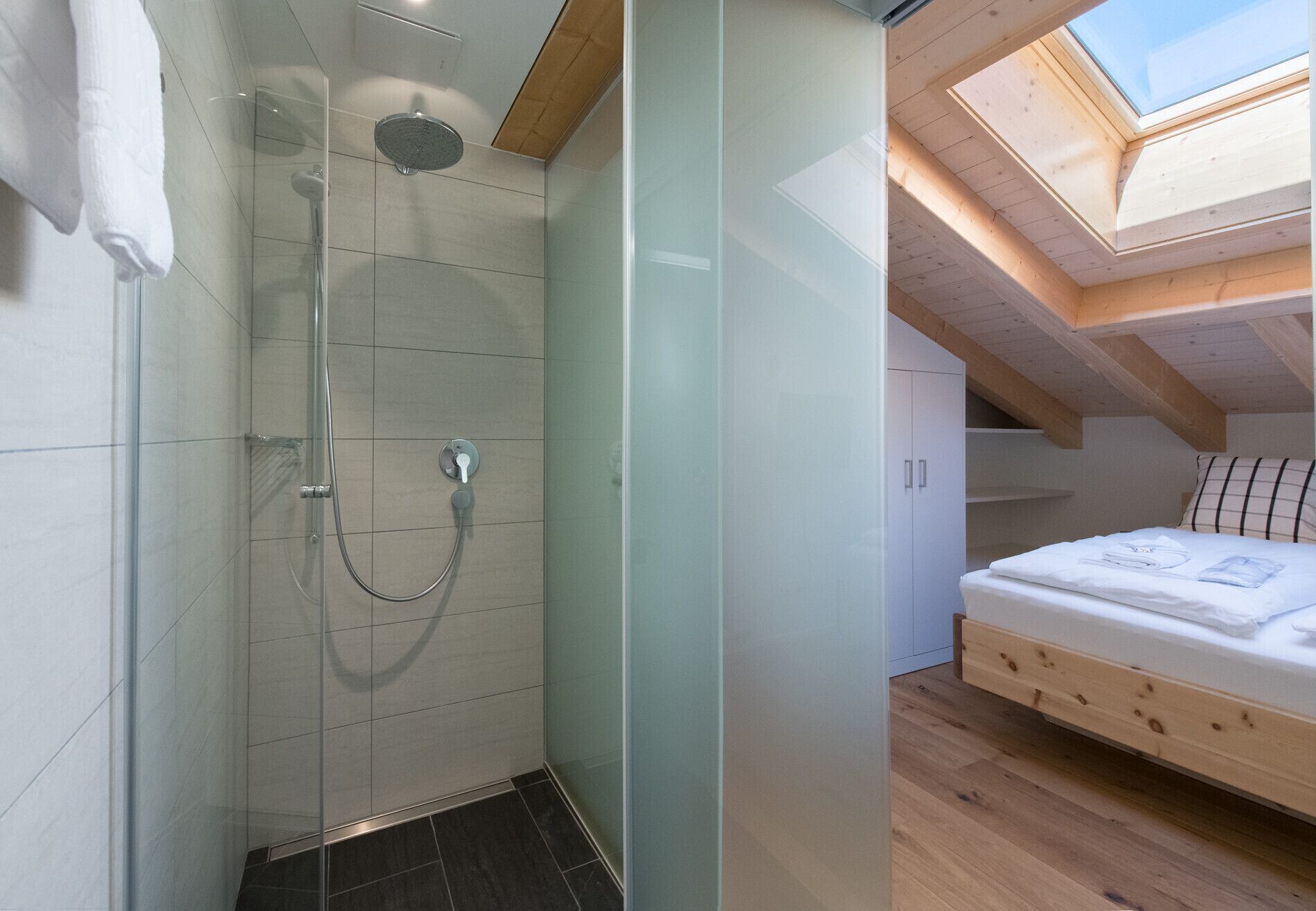 A modern bathroom with a glass shower enclosure and a skylight above a bedroom with a wooden bed frame.