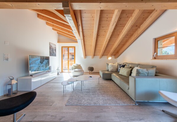 A modern living room with a high ceiling, wooden beams, a large flat-screen TV, a sofa, and a coffee table.