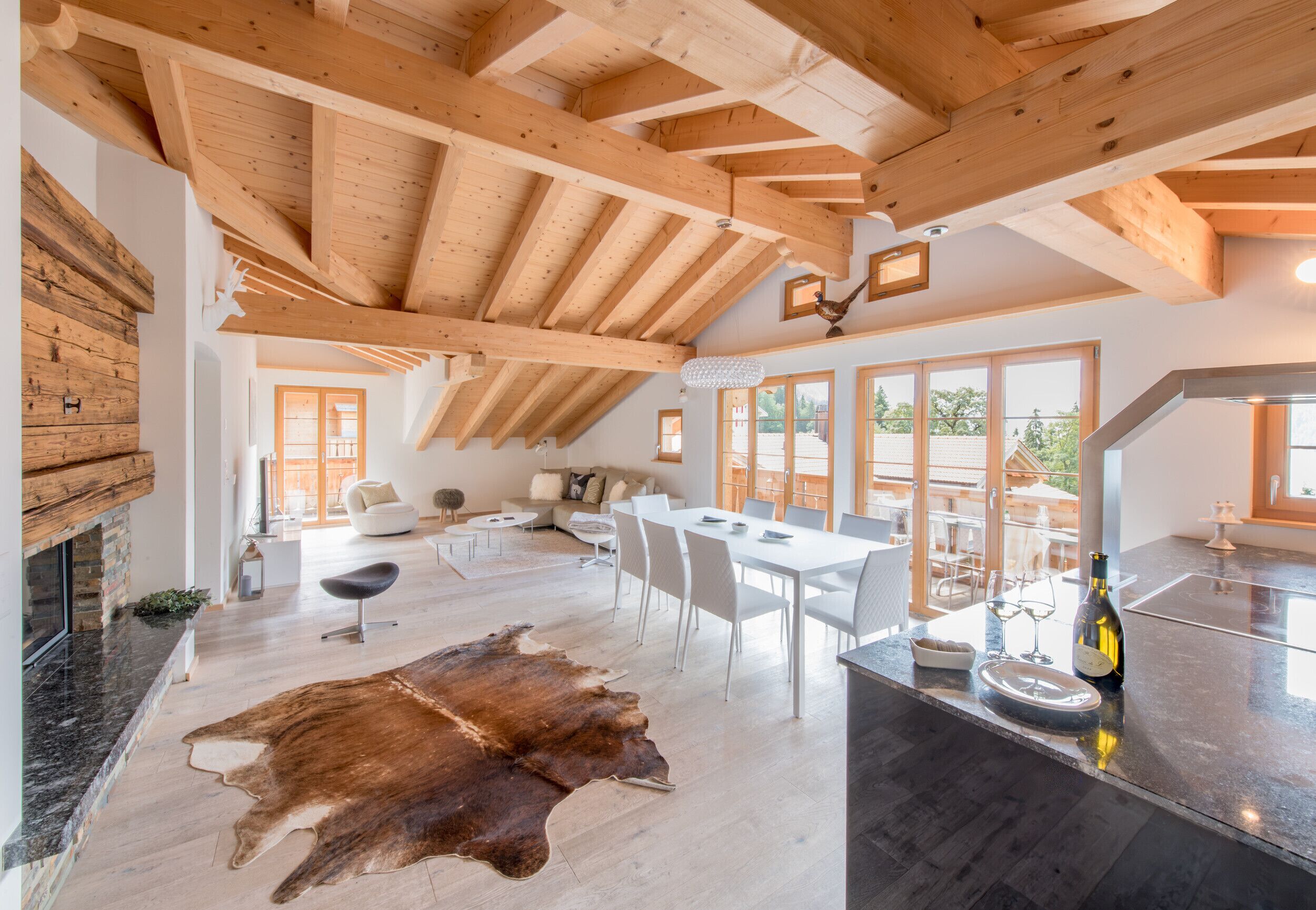 Amplia sala de estar con techo alto, vigas de madera, un gran tapete de cuero, una mesa de comedor con sillas y una zona de cocina con una botella de vino.