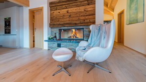 A modern living room with a stone fireplace, wooden ceiling beams, and a white armchair with a throw blanket.