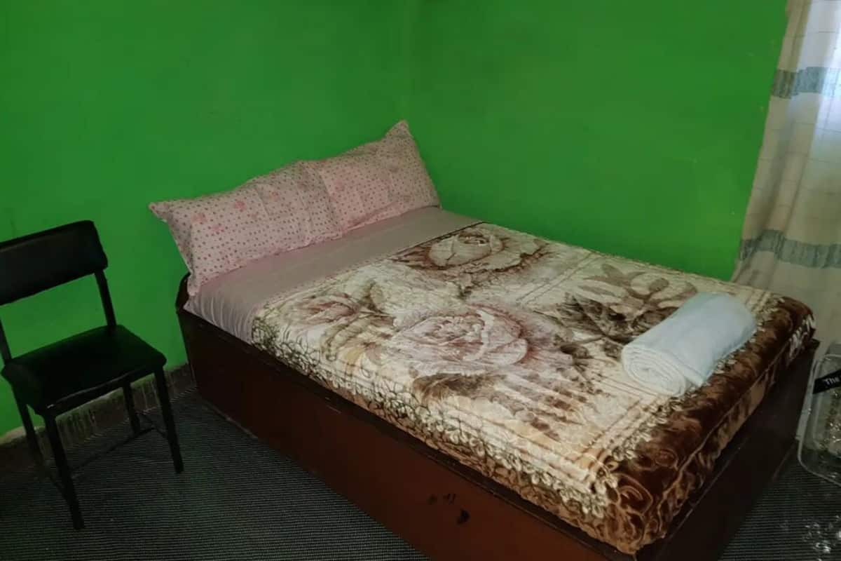 Un lit simple avec une couvre-lit à motifs floraux, un oreiller et une serviette enroulée. Une chaise noire est placée à gauche du lit.