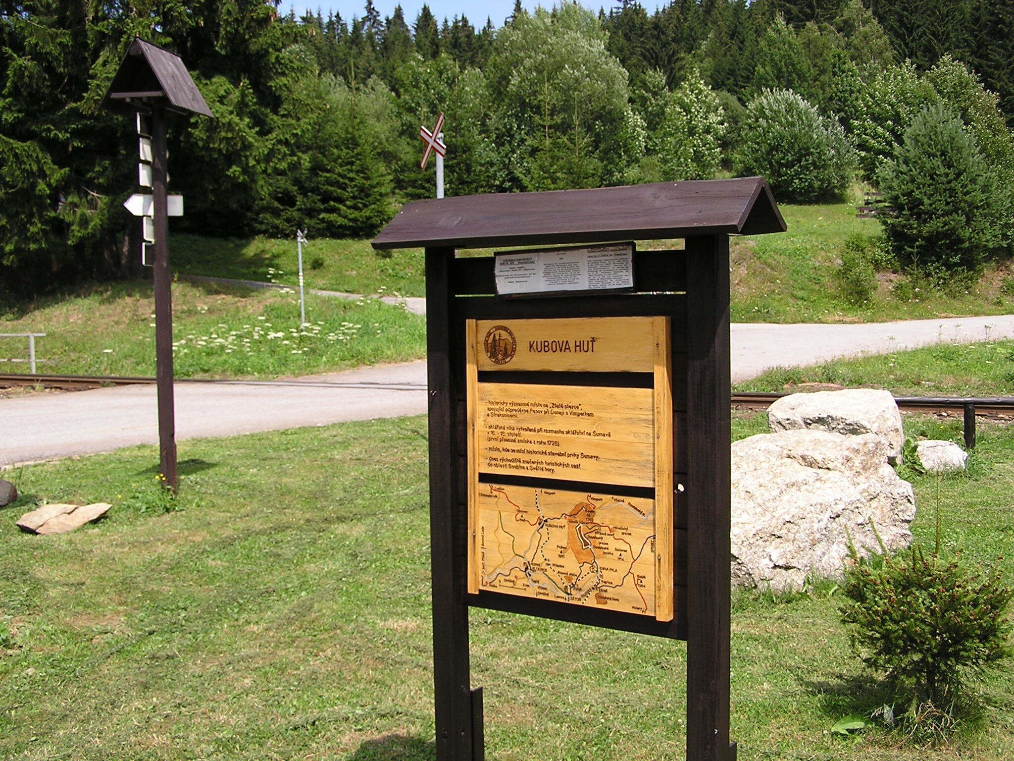 Ein hölzernes Schild mit einer Karte und Text, gelegen in einer grünen Umgebung mit einer Straße und Bäumen im Hintergrund.